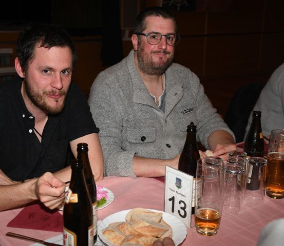 Zwei Männer sitzen an einem Tisch mit Essen und Bierflaschen. Der Mann auf der linken Seite trägt ein schwarzes Hemd und eine Brille. Der Mann auf der rechten Seite trägt einen grauen Pullover. Der Tisch hat ein Nummernschild 13, einen Teller mit Essen und Gläser Bier. Ein weiterer Mensch sitzt hinter ihnen.