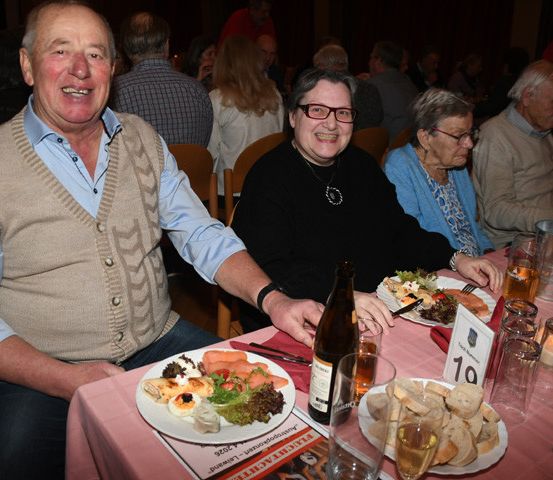 Eine Gruppe älterer Menschen sitzt an einem Tisch, lächelt und genießt ihr Essen. Sie sind von Tellern mit Essen, Gläsern und Weinflaschen umgeben. Der Tisch ist mit einer rosa Tischdecke bedeckt. Der Mann trägt eine Weste, während die Frau eine Brille trägt. Der Hintergrund ist unscharf, mit anderen sitzenden und stehenden Personen.