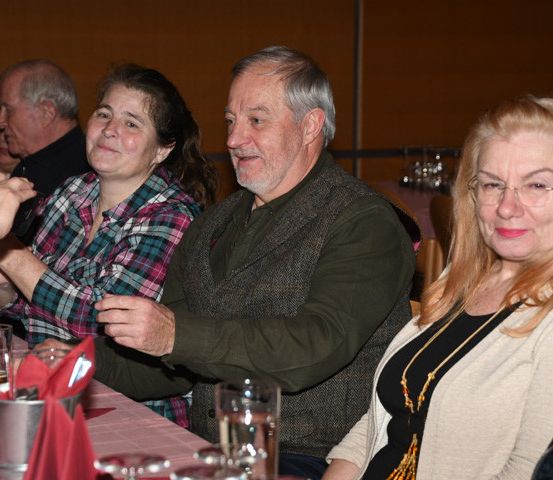Eine Gruppe von Menschen sitzt an einem Tisch, wahrscheinlich bei einem gesellschaftlichen Ereignis. Der Mann in der Mitte lächelt und hält etwas in den Händen. Die Frau auf der rechten Seite trägt eine Brille und eine Halskette. Der Tisch ist mit Gläsern und einem silbernen Becher besetzt.