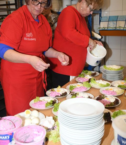 Zwei Frauen in roten Uniformen bereiten Gerichte auf einem Tisch zu, umgeben von Tellern und Schüsseln, in einer Küchenumgebung.
