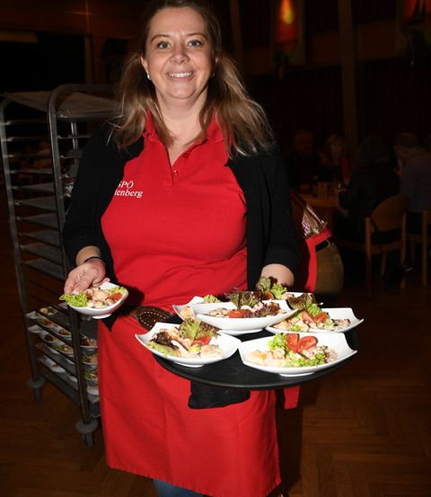 Eine Frau in einer roten Schürze hält einen Tablett mit Essensgerichten in einem Restaurant. Sie lächelt.