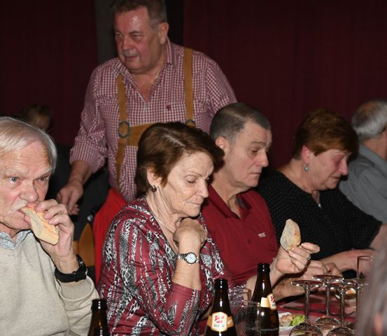 Eine Gruppe älterer Menschen sitzt an einem Tisch, isst, trinkt Bier und hält Essen. Flaschen Bier stehen auf dem Tisch, zusammen mit Weingläsern. Ein Mann in einem karierten Hemd und Hosenträgern steht hinter ihnen.