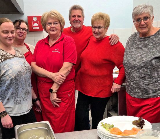 Eine Gruppe von Menschen, möglicherweise Freiwillige, steht in einer Küche. Einige tragen rote Schürzen. Vor ihnen steht ein Teller mit Essen.