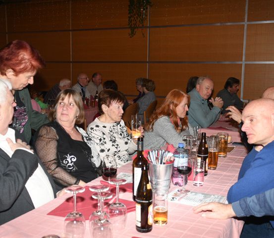 Eine Gruppe von Erwachsenen, sowohl Männer als auch Frauen, sitzt an einem Tisch, unterhält sich und genießt ihre Getränke. Der Tisch ist mit Weingläsern, Flaschen und einer rosa Tischdecke geschmückt.