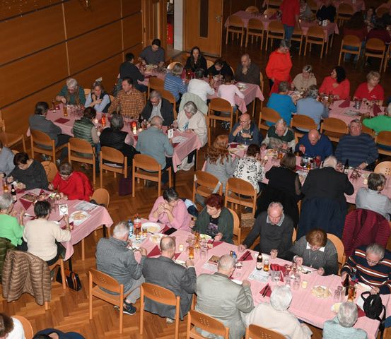 Ein großer Speisesaal mit mehreren Tischen und Stühlen ist mit vielen sitzenden und essenden Menschen gefüllt. Die Tische sind mit rosa Tischdecken bedeckt und haben Essen und Getränke darauf.