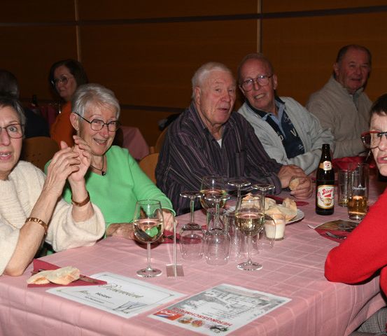 Mehrere ältere Erwachsene sitzen an einem Tisch, einige lächeln, in einem Restaurant. Sie haben Weingläser, Essen und Bierflaschen auf dem Tisch.