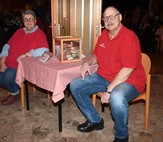 Zwei Personen sitzen an einem Tisch mit einer Schachtel darauf. Sie sind in einem Raum mit anderen Personen, die an Tischen sitzen.