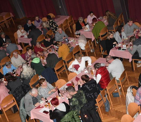 Viele Menschen speisen in einem Saal mit pinken Tischdecken. Ein Mensch steht neben einem Tisch mit dem Wort 'ART' darauf.