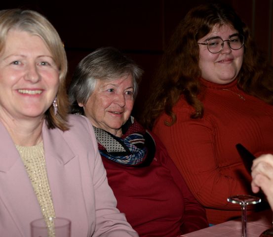Drei Frauen und ein Mann sitzen lächelnd an einem Tisch. Die Frauen tragen rote Oberteile und Schals, und der Mann hält Messer und Gabel. Gläser und Teller stehen auf dem Tisch.