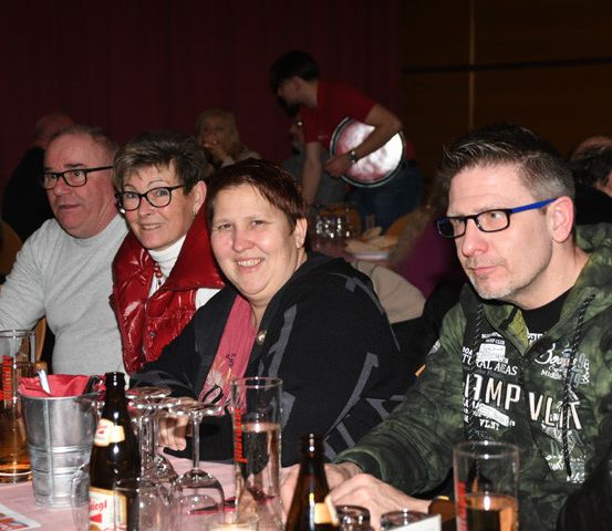 Eine Gruppe von Menschen sitzt an einem Tisch mit Gläsern und Flaschen, lächelt und schaut in die Kamera. Sie sind wahrscheinlich auf einer Party.