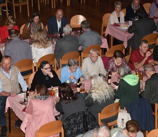 Eine Gruppe von Menschen diniert in einem großen Saal, mit Tischen, die mit rosa Tüchern bedeckt sind. Sie sitzen um die Tische herum, einige mit Flaschen und Gläsern. Der Raum ist aus Holz.