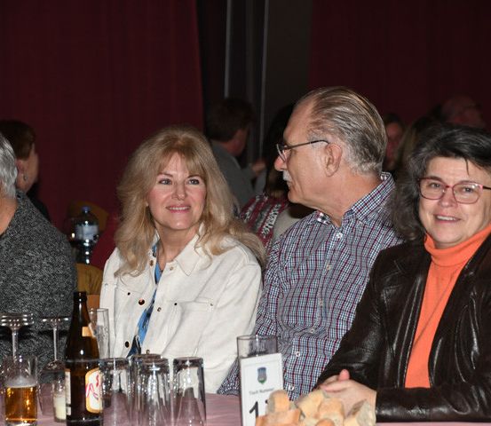 Mehrere Erwachsene sitzen an einem Tisch mit Weingläsern und Flaschen und lächeln für die Kamera. Rote Vorhänge im Hintergrund.