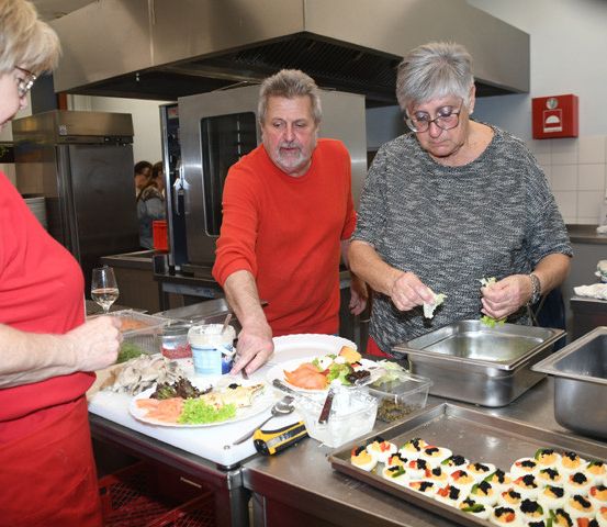 Drei Personen befinden sich in einer Küche und bereiten Essen zu. Eine Frau in einem roten Shirt hält einen Teller, ein Mann in einem roten Shirt hält einen Löffel und eine Frau in einem grauen Shirt bereitet einen Salat zu.