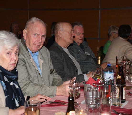 Mehrere ältere Menschen sitzen an einem langen Tisch und trinken aus Gläsern und Flaschen. Sie schauen auf etwas vor sich. Der Tisch hat Weingläser, Flaschen und einen Eimer. Die Wand hinter ihnen ist in einer warmen Farbe gestrichen.