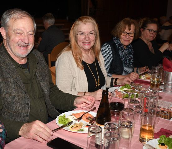 Eine Gruppe von Menschen sitzt an einem langen Esstisch. Sie lächeln und scheinen für ein Foto zu posieren. Sie halten Gabeln und Messer. Auf dem Tisch stehen Teller, Gläser und Flaschen. Der Tisch ist mit einer rosa Tischdecke bedeckt. Im Hintergrund sitzen noch weitere Menschen.