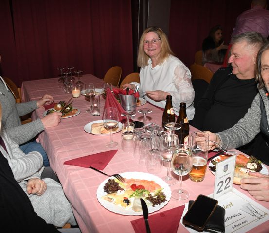 Eine Gruppe von Menschen sitzt an einem Tisch mit Essen und Getränken. Sie lächeln und scheinen sich zu amüsieren. Der Tisch ist mit Tellern, Gläsern und Servietten gedeckt.