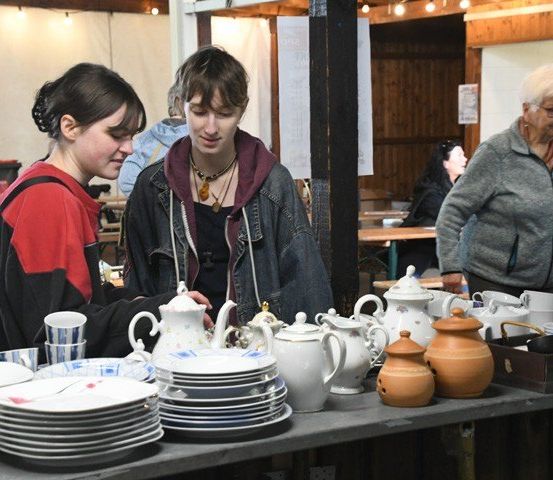 Zwei Frauen schauen Porzellan und Tee-Sets auf einem Tisch in einem Laden an. Andere Leute sind im Hintergrund.