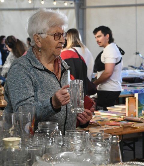 Eine ältere Frau hält einen klaren Glasbecher auf einem Markt mit vielen Gläsern und Büchern auf dem Tisch.