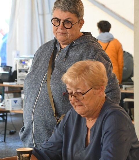 Zwei Frauen sitzen in einem Zelt an einem Tisch, eine trägt eine Brille und eine Jacke, die andere ein blaues Shirt und sieht ernsthaft aus. Ein Becher und ein Tablett mit Essen stehen auf dem Tisch.