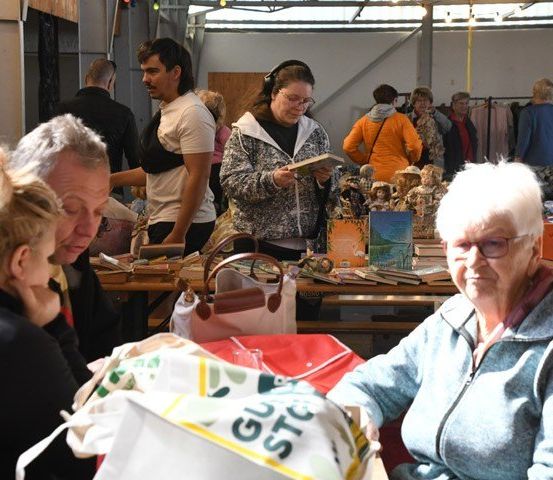 Eine Gruppe von Menschen in einem Raum mit Tischen voller Gegenstände. Eine Frau liest ein Buch und ein Mann spricht mit einer Frau.
