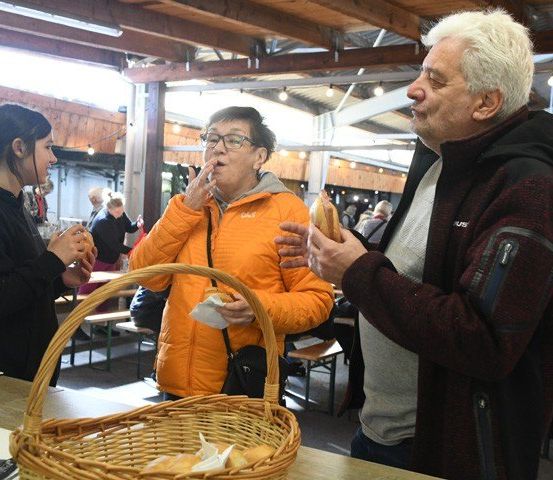 Eine Frau in einer orangefarbenen Jacke isst einen Sandwich auf einem Markt mit einem Mann und einer anderen Frau. Ein Korb mit Brot steht auf dem Tisch.