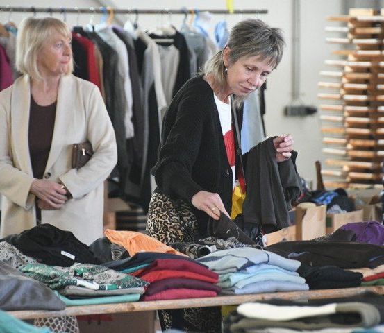 Zwei Frauen stehen vor einem Tisch mit gefalteter Kleidung in einem Geschäft. Eine Frau hält ein Kleidungsstück und die andere sieht es sich an. Hinter ihnen hängen Kleidungsstücke an Kleiderbügeln.