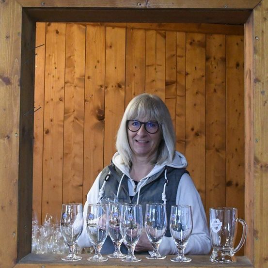 Eine Frau in einem Holzraum mit Gläsern und einem großen Krug. Sie lächelt und hält mehrere Gläser.