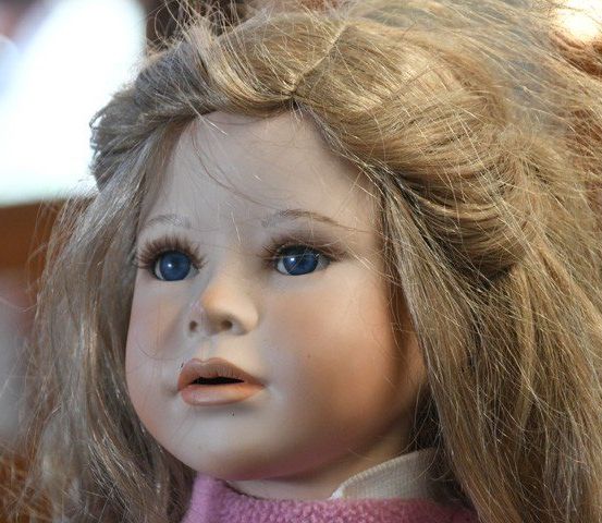 Eine Puppe mit langen blonden Haaren und blauen Augen trägt ein rosa Sweatshirt. Das Licht reflektiert auf dem Gesicht und den Haaren der Puppe.