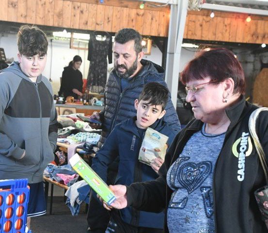 Eine Familie steht an einem Tisch mit einer Frau, die eine Schachtel hält. Zwei Jungen schauen sich die Schachtel an. Die Frau trägt eine Brille. Ein Mann in einem Mantel sieht die Jungen an.