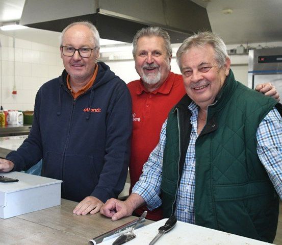 Drei Männer in einer Küche. Einer hält eine Box, ein anderer hat ein Messer und der dritte lächelt.