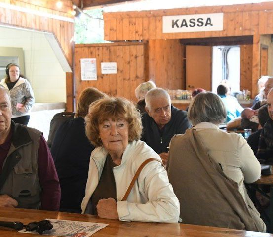Mehrere ältere Menschen sitzen an Tischen in einem Restaurant. Ein Schild darüber steht Kassa. Eine Frau im Hintergrund trägt Kopfhörer.