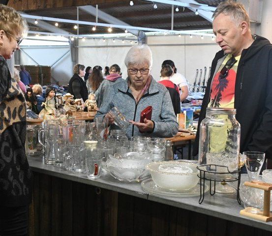 Mehrere Menschen kaufen auf einem Markt mit verschiedenen Artikeln ein. Eine ältere Frau betrachtet Glaswaren. Ein Mann in einem Kapuzenpullover schaut sich einen Krug an. Dahinter stöbern Leute durch andere Produkte.