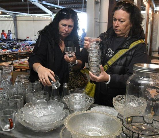 Zwei Frauen betrachten Glaswaren in einem Indoor-Markt. Eine Frau hält ein Glas Wasser und die andere hält einen Stapel Gläser.