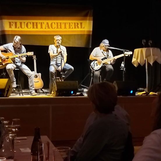 Drei Musiker spielen auf der Bühne mit Gitarren, einer singt in ein Mikrofon. Ein Banner mit dem Text Fluchtachterl im Hintergrund. Ein Tisch mit Weingläsern und einer Flasche im Vordergrund.