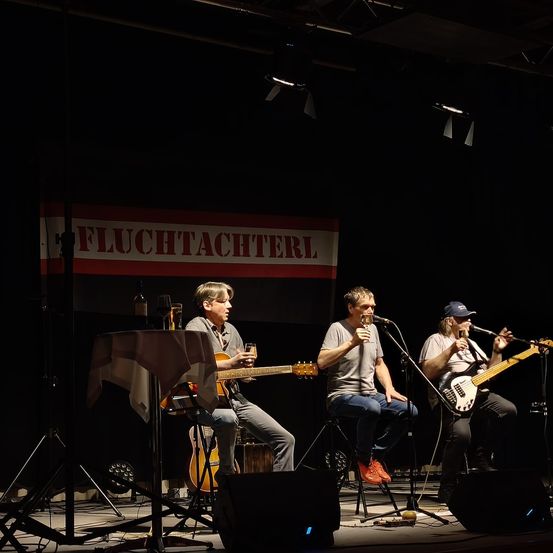 Drei Musiker stehen auf der Bühne unter einem Schild, das Fluchtachterl sagt. Sie sitzen auf Hockern und spielen Gitarren und Bass. Einer von ihnen hält ein Glas.