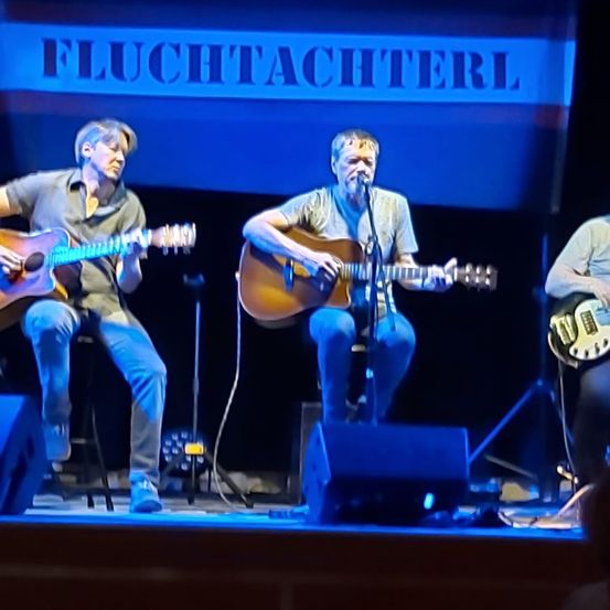 Drei Männer spielen auf der Bühne und spielen Gitarre. Einer in der Mitte singt in ein Mikrofon. Ein Schild oben sagt Fluchtachterl.