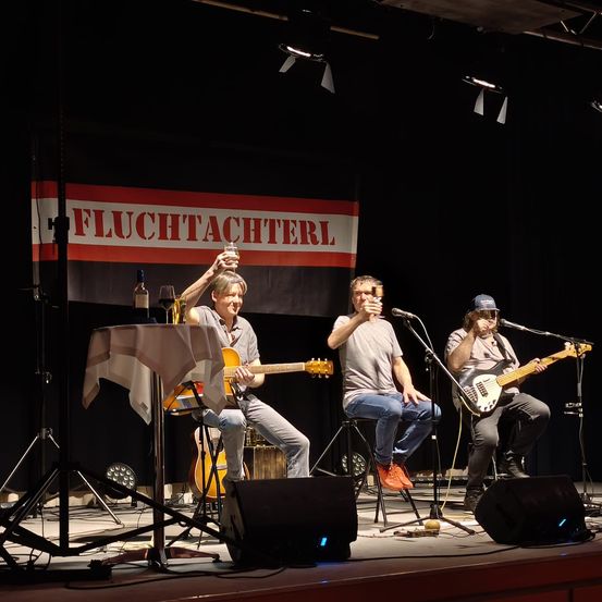 Drei Männer spielen auf der Bühne mit Gitarren, ein Banner mit dem Text Fluchtachterl im Hintergrund und Flaschen und Mikrofone vor ihnen.