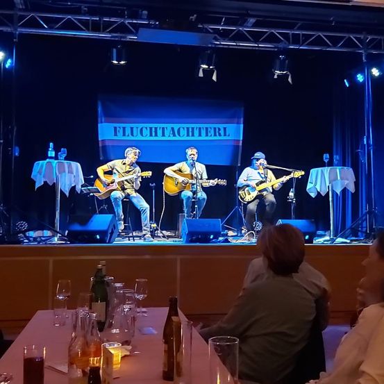 Drei Musiker spielen auf der Bühne mit akustischen Gitarren. Hinter ihnen befindet sich ein Banner mit der Aufschrift 'FLUCHTACHTERL'. Unter der Bühne schaut ein Publikum zu, mit Flaschen und Gläsern auf dem Tisch davor.