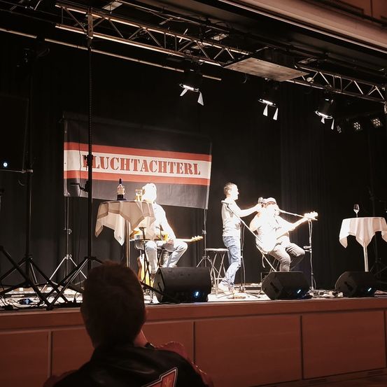 Eine Band spielt auf einer Bühne mit zwei Gitarristen, einer mit Mikrofon, und einem Banner mit der Aufschrift 'Fluchtachterl'. Scheinwerfer und Lautsprecher sind aufgestellt, und ein Publikum ist anwesend.