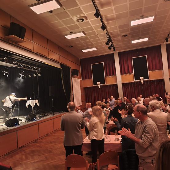 Ein Musiker spielt Gitarre auf einer Bühne in einem Saal mit klatschende Zuschauer. Die Bühne hat einen schwarzen Hintergrund und Lautsprecher. Der Saal hat Holzböden, rote Vorhänge und Deckenleuchten.