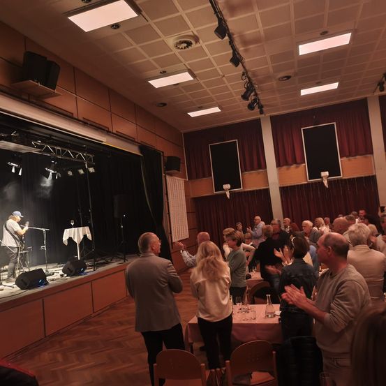Eine Indoor-Veranstaltung mit einem Künstler auf der Bühne, umgeben von einem Publikum, das an Tischen sitzt, unter einer Decke mit Lichtern und einer Bühnenkulisse.