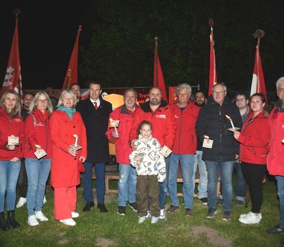 Eine Gruppe von Menschen in roten Jacken hält Kerzen vor einem Banner. Sie posieren für ein Foto bei Nacht.