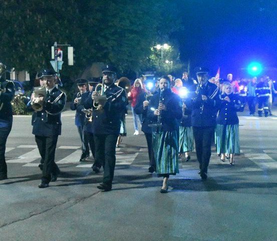 Eine Band in Uniform spielt nachts auf einer Stadtstraße Instrumente, mit Zuschauern und Rettungsfahrzeugen im Hintergrund.