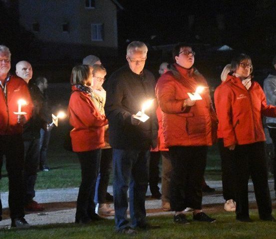 Eine Gruppe von Menschen in roten Jacken steht in einer Reihe und hält Kerzen bei Nacht, möglicherweise bei einer Gedenkfeier.