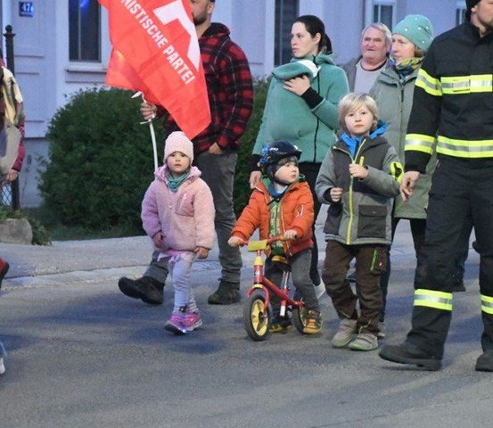 Eine Gruppe von Kindern und Erwachsenen geht auf einer Straße, ein Kind fährt Fahrrad, ein Feuerwehrmann folgt, ein Mann hält eine rote Fahne.