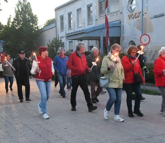 Eine Gruppe von Menschen in roten Jacken geht auf einer Straße und hält brennende Kerzen. Sie stehen vor einem Gebäude mit einer Wandmalerei und einer Fahne.