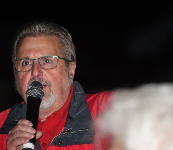 Ein Mann mit grauem Haar trägt eine Brille und eine rote Jacke. Er spricht in ein Mikrofon.