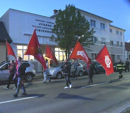 Eine Gruppe von Personen, die mit roten Fahnen vor einem Jugend-gelabelten Gebäude die Straße hinunter marschieren.
