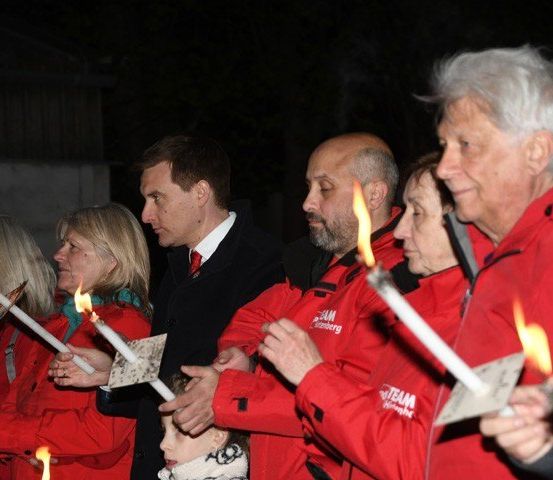 Eine Gruppe von Menschen in roten Jacken hält brennende Kerzen bei Nacht; ein erwachsener Mann in einem Anzug steht unter ihnen.