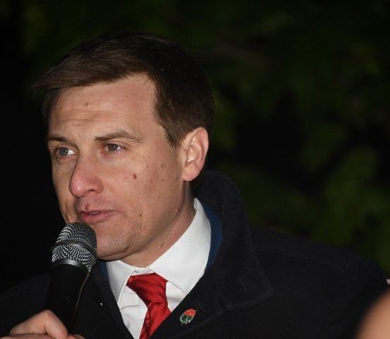 Ein Mann in einem schwarzen Mantel und roter Krawatte spricht in ein Mikrofon in einer schwach beleuchteten Außenumgebung.